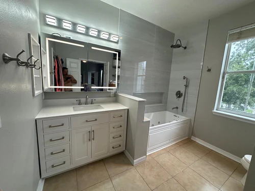 The finished Celebration bathroom from the vanity side, with a lit mirror, gray porcelain tile, and the Jacuzzi tub under the window