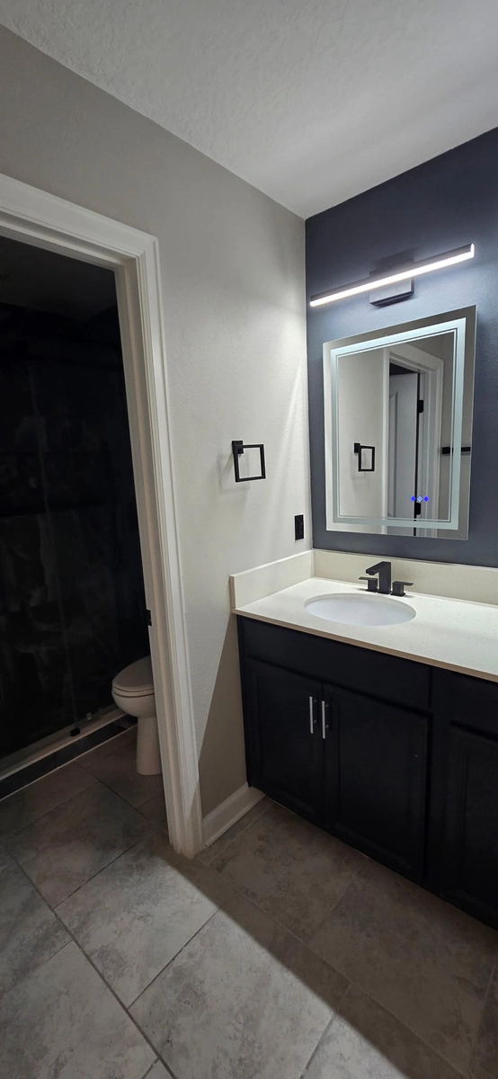 The new sink side of the Kissimmee bathroom, with a dark vanity, a lit mirror on a navy accent wall, an LED light bar, and matte black fixtures