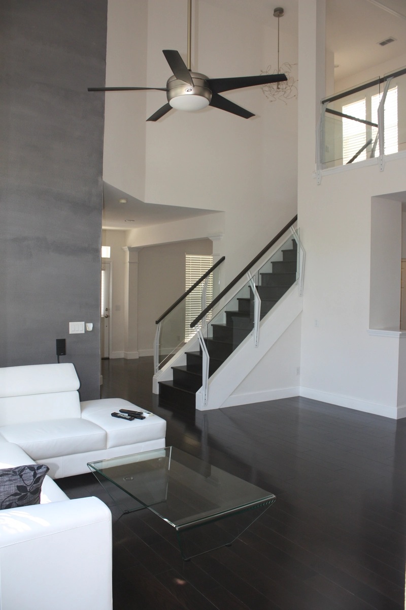 Finished Celebration living area with dark hardwood floors, a gray accent wall, a glass panel stair railing, and a modern fan hung in the two story ceiling